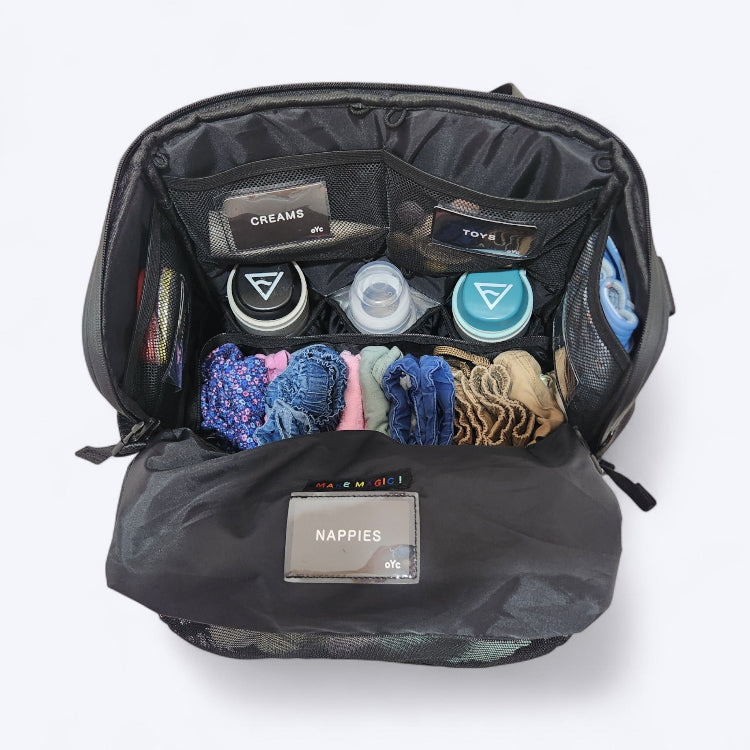 OUT YNDR CLUB Medley nappy bag, black colour, top open view showing lots of pockets and compartments filled with baby bottles, creams, clothes and nappies. Organised layout.